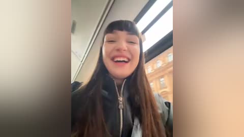 Media: A video of a young Asian woman with long brown hair, wearing a black jacket, laughing joyfully. She is inside a train, with large windows showing a cityscape outside.