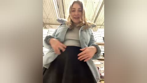 Media: Video of a smiling woman with shoulder-length blonde hair, wearing a light blue jacket over a grey top and a black skirt, standing in a warehouse with metal beams and shelves in the background.