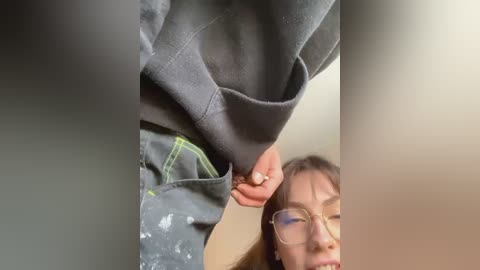 Media: Video of a woman with glasses, wearing a grey sweatshirt with a hood and black jeans, sitting indoors with a neutral background.