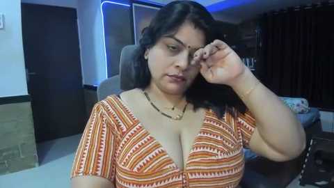 Media: Video of a South Asian woman with medium brown skin, wearing a low-cut orange patterned top, adjusting her glasses in a dimly lit room with dark curtains and a white wall.