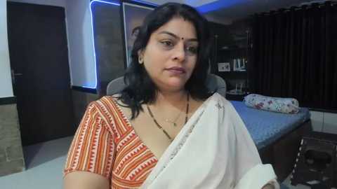 Media: Video of a South Asian woman with medium skin tone and dark hair, wearing an orange and white patterned sari, in a modern bedroom with a bed, dark curtains, and a closed door.