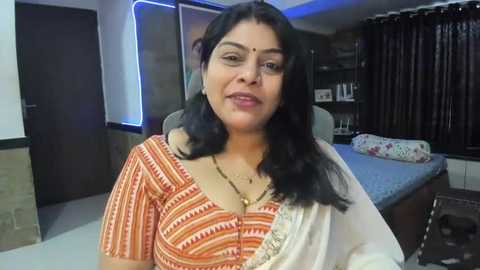 Media: Video of an Indian woman with long black hair, wearing an orange and white striped blouse, sitting in a modern bedroom with a bed and dark curtains.