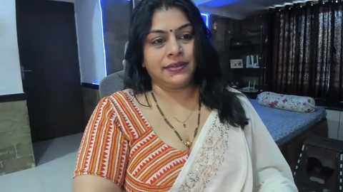 Media: Video of an Indian woman with medium skin tone, long black hair, and traditional attire, sitting in a modern bedroom with a bed, nightstand, and shelves.