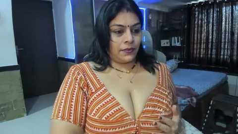 Media: A video of a middle-aged South Asian woman with dark hair and medium skin tone, wearing a low-cut orange patterned dress, sitting in a modest, cluttered bedroom with a bed and bookshelf.