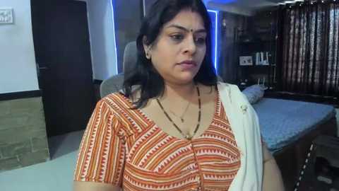 Media: Video of an Indian woman with medium skin tone and black hair, wearing a traditional orange and white striped saree, seated in a modern living room with dark wooden furniture and blue lighting.