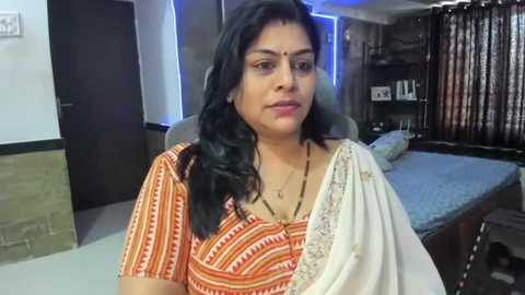 Media: Video of an Indian woman in a white sari with orange and white geometric patterns, sitting on a bed in a modern, cozy bedroom with dark wooden furniture, blue lighting, and a neatly made bed.