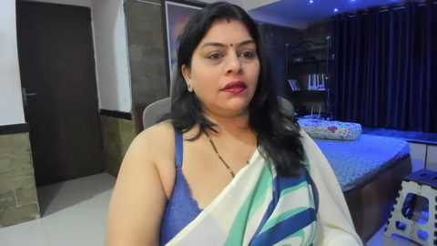 Media: Video of a South Asian woman with medium skin tone, long black hair, wearing a white and blue saree, sitting in a modern bedroom with a bed, curtains, and bookshelf.