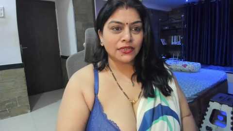 Media: Video of a South Asian woman with medium brown skin, black hair, and a medium build, wearing a blue bra and a white and blue shawl, sitting on a grey chair in a modern bedroom with a bed, bookshelf, and dark curtains.