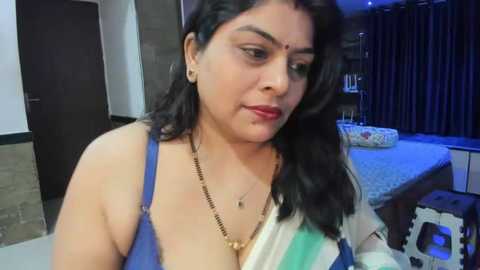 Media: Video of a middle-aged South Asian woman with medium skin tone, wearing a blue and white striped top, sitting in a modern, dimly lit bedroom with dark curtains.