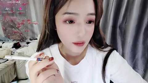 Media: Video of a young Asian woman with fair skin, long dark hair, and light pink makeup, applying lipstick with a silver applicator, sitting in a messy room with a pink cherry blossom background.