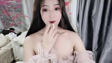 Media: Video of an East Asian woman with long black hair, fair skin, and pink lipstick, biting her finger, wearing a sheer, off-shoulder, light pink dress, in a room with floral wallpaper and draped curtains.