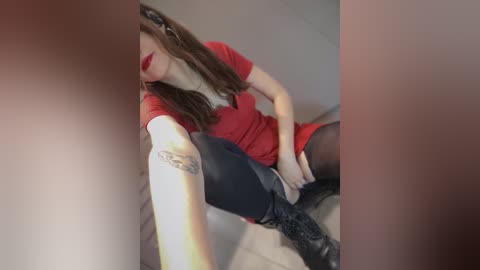 Video of a fair-skinned woman with long brown hair, red lipstick, wearing a red top, black leggings, and black boots, sitting with legs spread, in a dimly lit room.