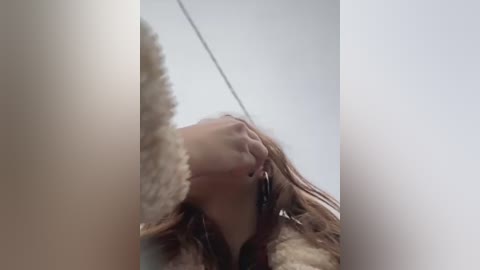 Video of a woman with light skin and long brown hair, wearing a fur coat, standing in front of a mirror, touching her face. The background is blurred.