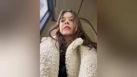 Media: A video of a young woman with long, wavy brown hair, wearing a fluffy beige blanket, seated inside a train car with beige seats and a window visible in the background.