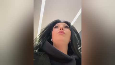 Media: Video of a woman with long, straight black hair, wearing a black coat and pink lipstick, standing in a modern, minimalist room with white walls and recessed ceiling lights.