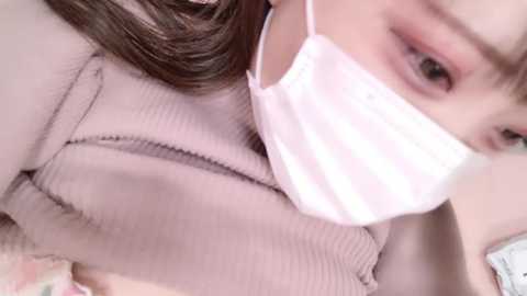 Media: Video of an Asian woman with fair skin, wearing a beige ribbed sweater, a white face mask, and brown hair, seen from a close-up angle, indoors.