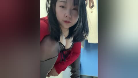 Media: A video of a young East Asian woman with straight black hair, wearing a red cardigan over a black lace bra, in a brightly lit room with blue walls and white tiles.