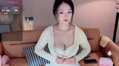 Media: Video of an East Asian woman with fair skin and dark hair, wearing a white blouse and cardigan, sitting on a beige couch, surrounded by pink and black patterned pillows.