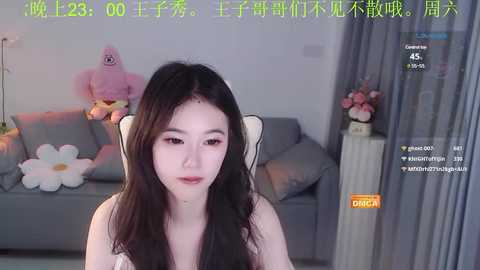 Media: A video of a young Asian woman with long black hair, fair skin, and a slender physique, seated in a modern, minimalist living room with a gray sofa, plush toys, and floral decor.