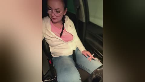 Media: A video of a woman with glasses and dark hair, wearing a white cardigan over a pink top and blue jeans, seated in a car, focusing on her phone.