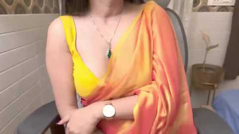 Media: Video of a woman in a vibrant yellow and orange saree, seated in a modern room with white tiled walls and a potted plant.