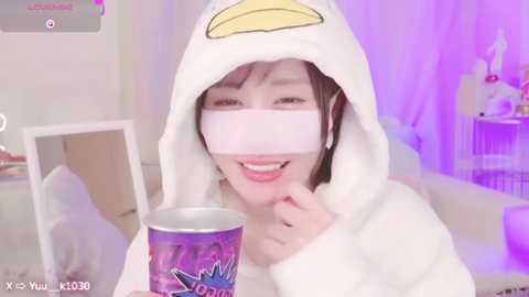 Media: Video of a young East Asian woman wearing a white, plush duck hooded robe and a white face mask, smiling, holding a purple cup. Background features a white room with a mirror, purple lighting, and a shelf with items.