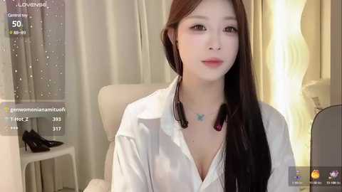 Media: A video of a young East Asian woman with long, straight black hair, wearing a white blouse with a butterfly necklace, sitting indoors with beige curtains.