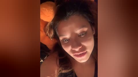 Media: Video of a young Asian woman with long black hair, light skin, and dark eyes, lying on a bed with an orange teddy bear and a dark blanket.