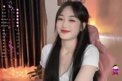 Media: Video of an East Asian woman with long black hair, fair skin, and red lipstick, smiling, wearing a white top, seated in a pink chair against a dimly lit room with curtains.