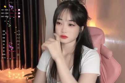 Media: Video of a young East Asian woman with long black hair, wearing a white t-shirt, sitting on a pink chair, looking contemplative. Background includes a lit window and a potted plant.