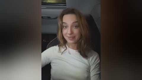 Media: A video of a smiling Caucasian woman with light brown hair, wearing a white long-sleeve shirt, seated in a car with dark interior.