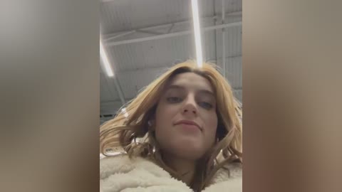 Media: A video of a young woman with long, wavy, light brown hair, wearing a fluffy, cream-colored sweater, standing in a dimly lit, industrial room with exposed metal beams and fluorescent lights.