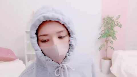 Media: Video of a young Asian woman with light skin, wearing a light blue hooded sweatshirt and a white surgical mask, standing indoors with a white wall and potted plant in the background.