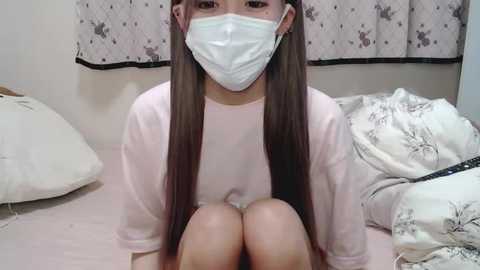 Media: A video of an Asian woman with long brown hair, wearing a white surgical mask, pink long-sleeve shirt, and white shorts, sitting on a bed with white sheets and floral-patterned pillows, in a room with light-colored curtains.