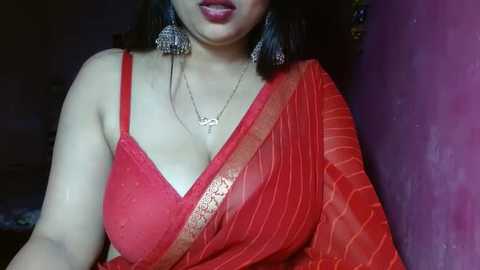 Media: Video of a South Asian woman with light brown skin, wearing a red, sheer sari with gold trim, and a matching bra, against a dark purple wall. She has shoulder-length dark hair and wears large, dangling earrings.