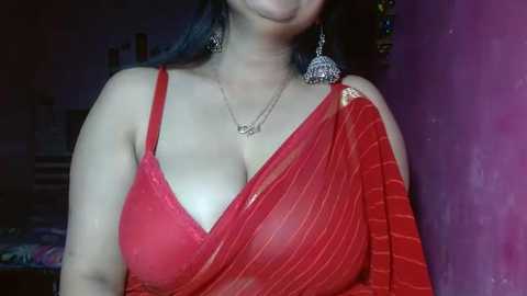 Media: Video of a woman with medium skin tone, wearing a sheer red saree, showing cleavage, large silver earrings, and a necklace. Background features a purple wall and indistinct objects.