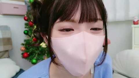 Media: Video of a young Asian woman with light skin, straight brown hair in pigtails, wearing a pink face mask and light blue scrubs, standing in a brightly lit room with a Christmas tree and presents in the background.