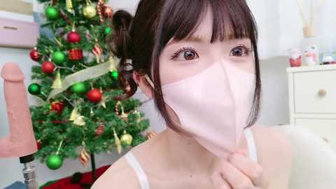 Media: Video of an Asian woman with fair skin, brown hair in buns, wearing a pink face mask, holding a realistic pink dildo in a festive room with a decorated Christmas tree.