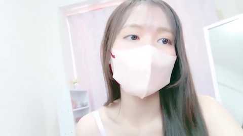 Media: Video of an East Asian woman with long, straight black hair, wearing a white face mask, pink earbuds, and a white tank top. Background features a white room with pastel decor and a mirror.