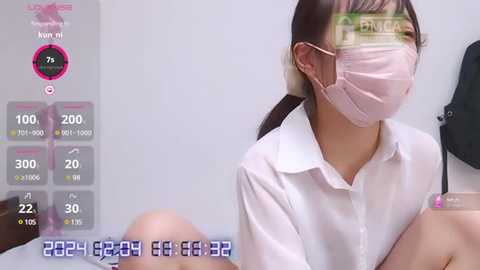 Media: A video of an East Asian woman with light skin and straight black hair, wearing a white shirt, pink surgical mask, and gloves, sitting indoors, blurred in the background.