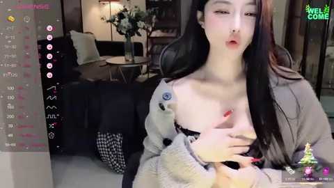 Media: Video of a young East Asian woman with long black hair, fair skin, and red lipstick, topless in a gray cardigan, sitting in a modern living room.