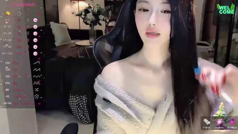 Media: Video of an Asian woman with long black hair, fair skin, and a slender physique, wearing a beige off-shoulder sweater. She sits on a black office chair in a dimly lit room with a TV screen displaying social media icons.