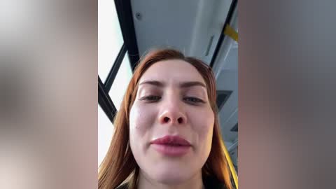 Media: A close-up video of a young woman with fair skin and long red hair, wearing minimal makeup, captured from a low angle. The background shows an interior with black-framed windows and a yellow object, possibly a piece of furniture or equipment.