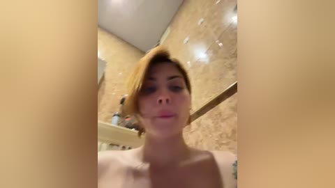 Media: Video of a light-skinned woman with short, blonde hair in a bathroom. She's topless, with visible breasts. The background features a beige-tiled wall and a blurred shelf with toiletries. The image is slightly blurry.