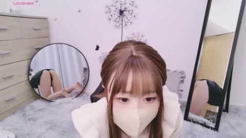 Media: Video of a young Asian woman with long brown hair, wearing a white mask, in a bedroom with white walls, a large mirror, and a grey dresser.