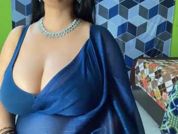 Media: Video of a woman with medium skin tone and black hair, wearing a deep blue, low-cut satin dress that accentuates her cleavage, against a backdrop of geometric patterned wallpaper and a colorful quilt.