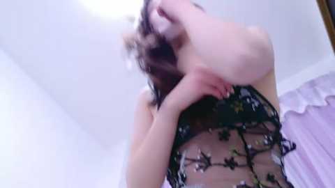 Media: Video of a pale-skinned woman with dark hair, wearing a sheer black lace bodysuit with floral embroidery, in a dimly lit bedroom with white walls and a lavender bedspread.