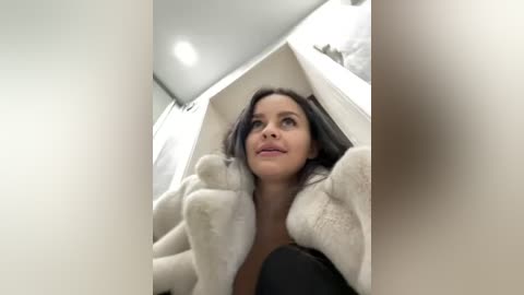 Media: Video of a smiling woman with long black hair, wearing a beige furry coat, inside a white doghouse.