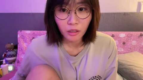 Media: Video of an Asian woman with straight shoulder-length brown hair, wearing round glasses, and a light gray t-shirt, sitting on a pink polka-dotted bedspread.