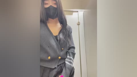 Media: A video of an Asian woman with long black hair, wearing a black face mask, grey ribbed blazer with gold buttons, and black skirt, standing in a dimly lit hallway.